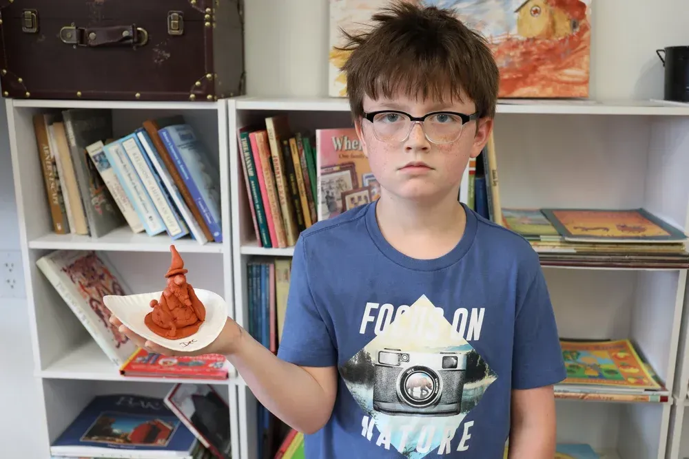 Boy holding a small plate with a red sculpture, standing in front of a bookshelf and display shelf.