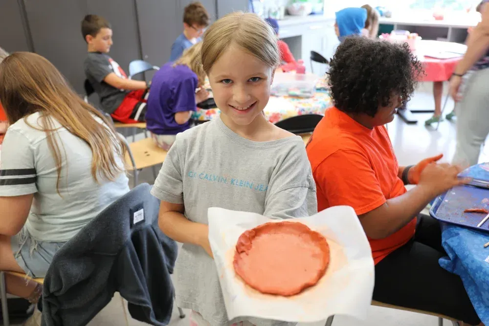 Child smiling in a classroom, holding a paper plate with an orange-painted circle artwork.