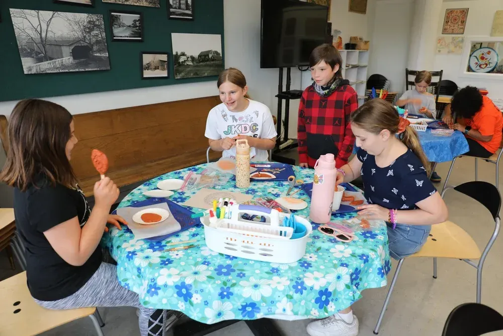People painting at a table with blue tablecloth in a bright classroom or craft room.