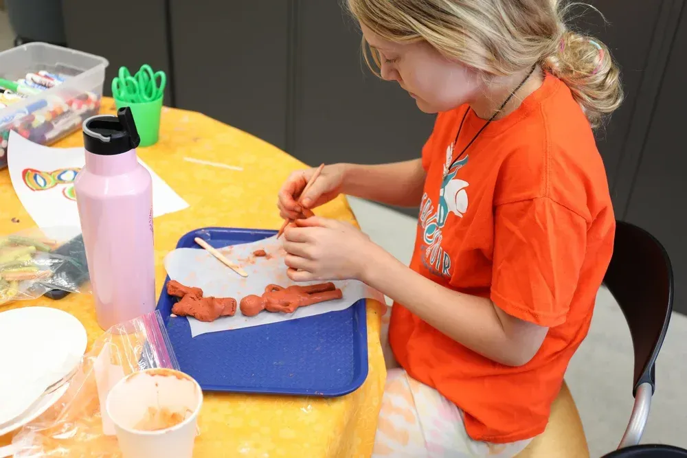 Child in orange shirt shaping clay at a table with tools, water bottle, and a blue tray