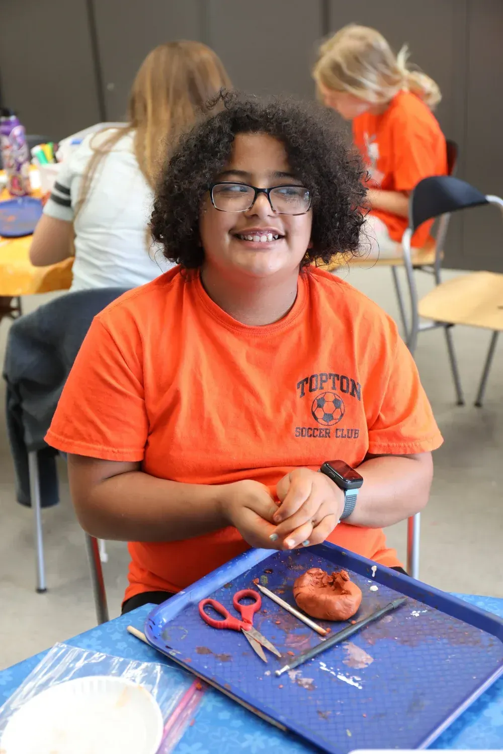 Student in orange shirt smiles at a craft table with scissors, a small item, and a blue placemat.