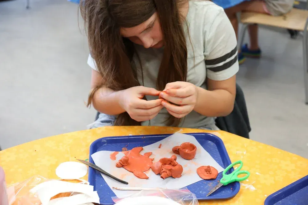 Person crafting with orange clay at a table, using tools on a blue tray.
