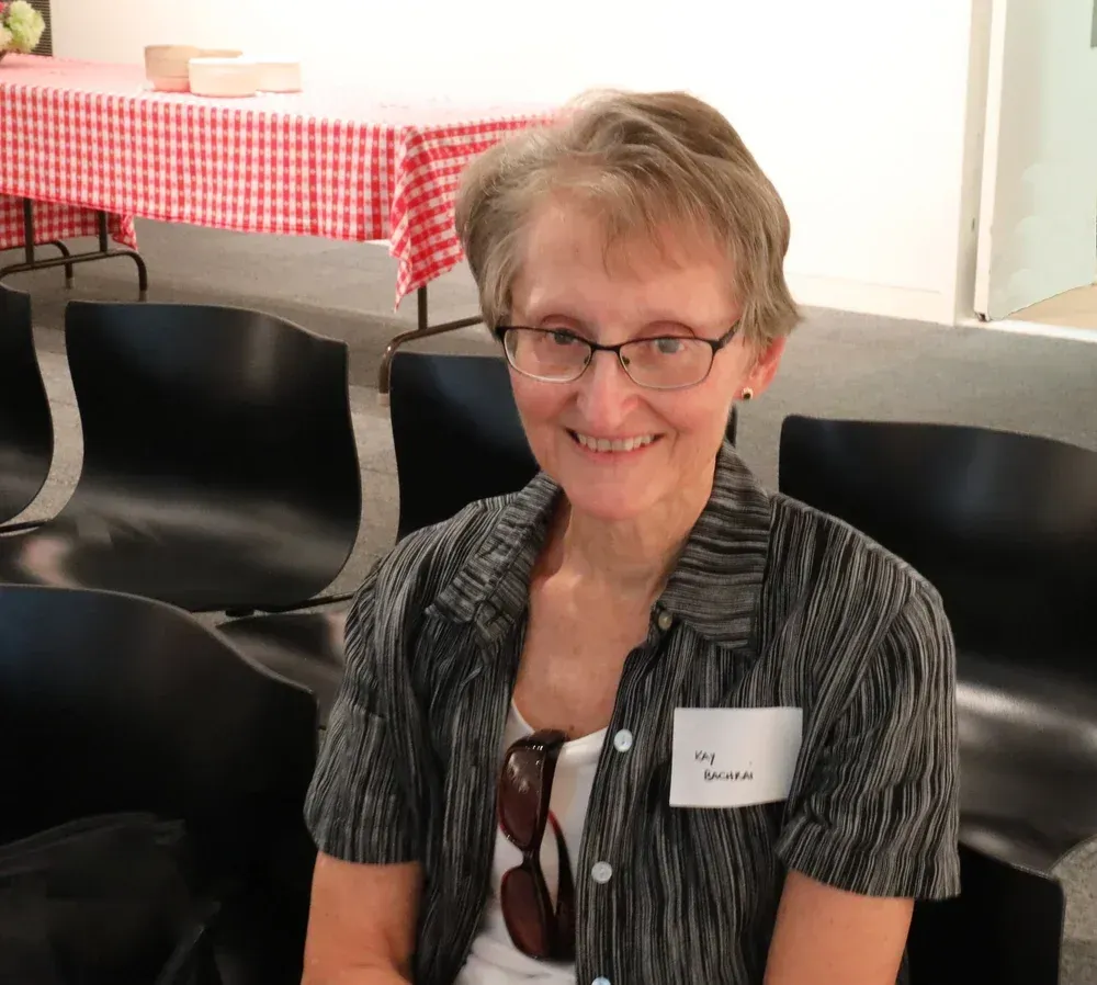 Smiling person seated indoors beside black chairs and a red-checkered tablecloth, wearing a name badge.