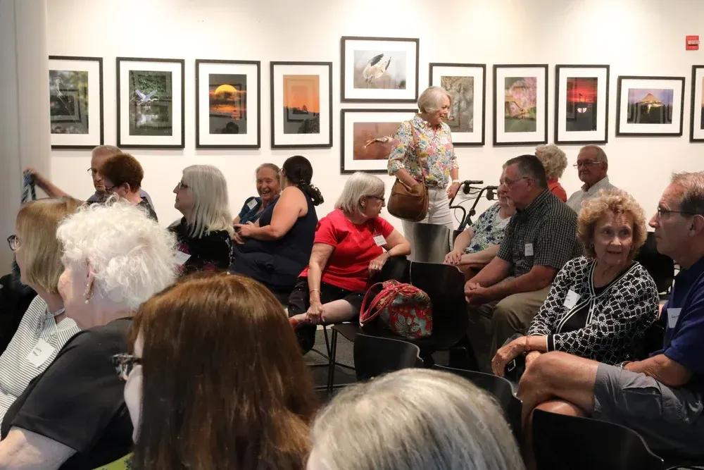 Audience seated in an art gallery for a presentation, with framed paintings on the wall.