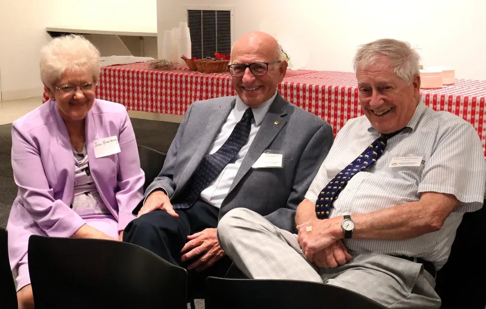 Three smiling people seated indoors, wearing name tags and formal attire, in front of a red checkered tablecloth.