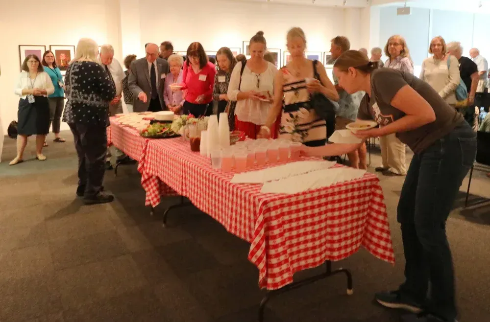 People serving food at a buffet table with red checkered cloth in a bright indoor room