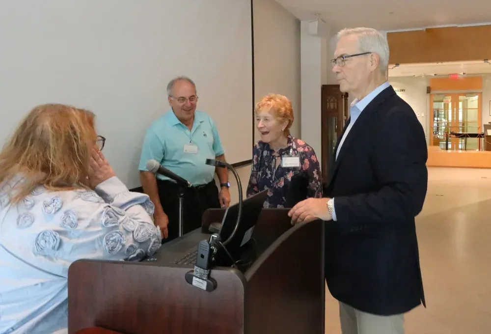 People gathered around a podium in a bright lobby, speaking and listening during an event.