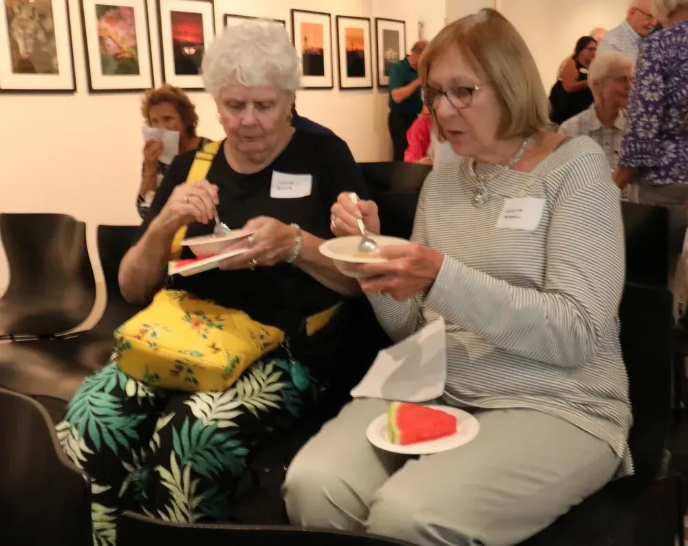 Two people seated indoors eating dessert from small bowls, with framed art on the wall behind them.