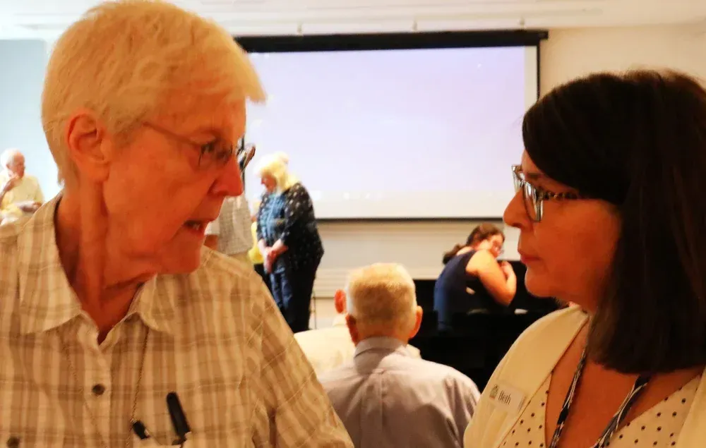 Two people talking at a conference, with a presentation screen in the background.