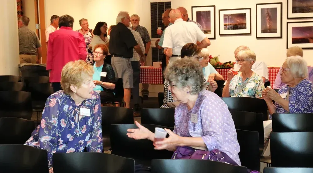 People chatting in a lounge, with framed photos on the wall and black chairs in the foreground.