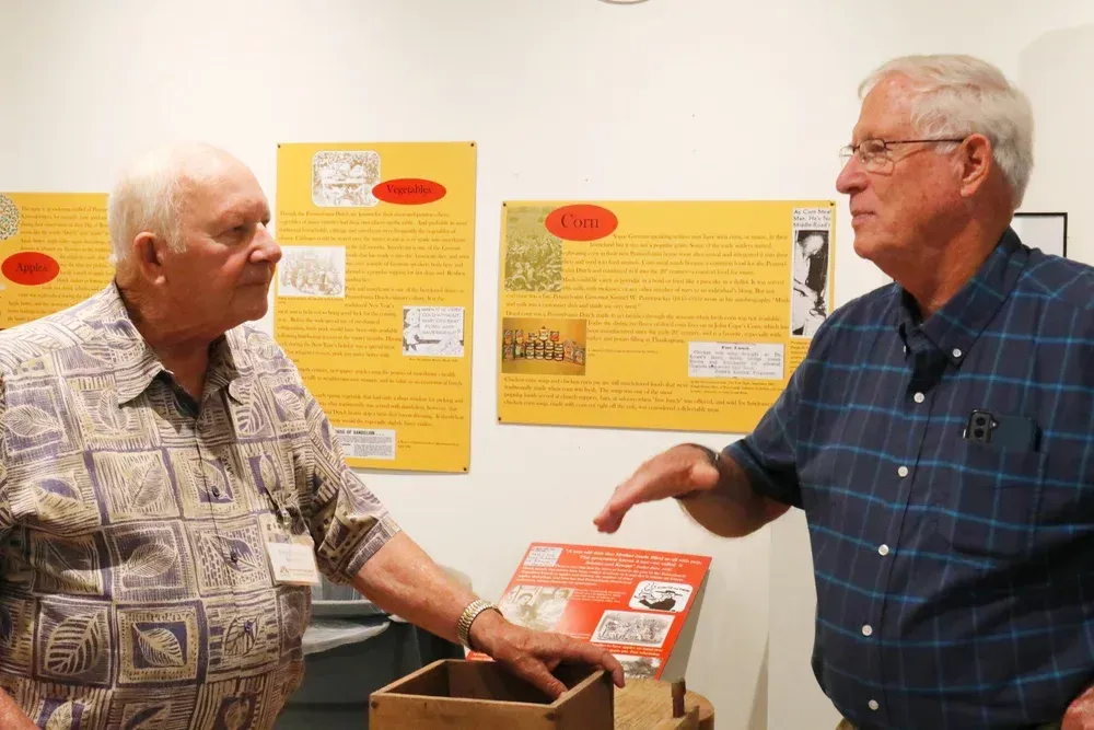 Two men talking beside display boards, one gesturing toward an orange poster in a bright room.