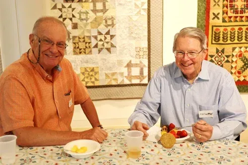 Two smiling men sitting at a quilt-covered table with cups and snacks in a cozy room
