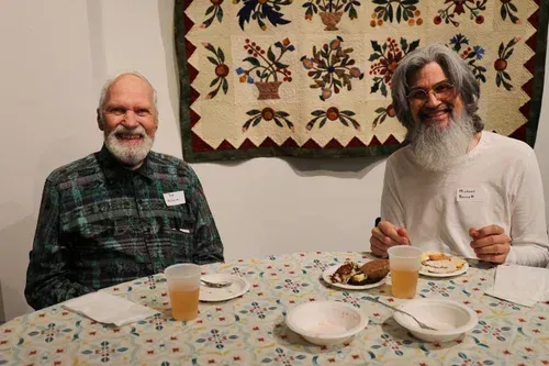 Two smiling men seated at a dining table with drinks and plates, indoors under a floral wall hanging