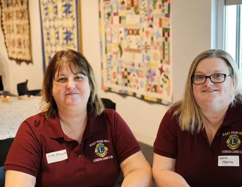 Two women in maroon polos sit in a room with colorful quilts displayed behind them.