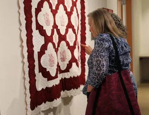 Woman in blue patterned top and red skirt viewing a red-and-white quilt on a wall display.
