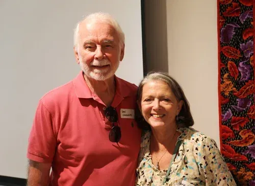 Two smiling people standing indoors, one in a red shirt and one in a floral blouse.