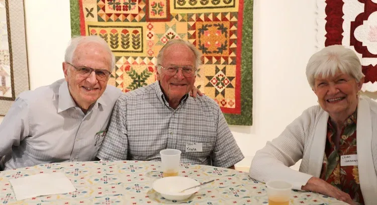 Three smiling people seated at a table in a bright room, with a colorful quilt hanging behind them.