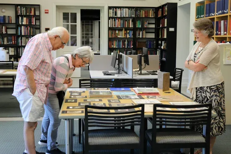 Three people examining books at a library table with a computer screen nearby