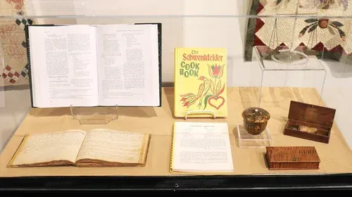 Museum display of open books, a yellow book, papers, and small historical objects in glass cases