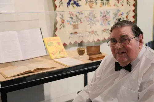 Man in glasses and a white shirt sits by a piano with sheet music and books in a bright room