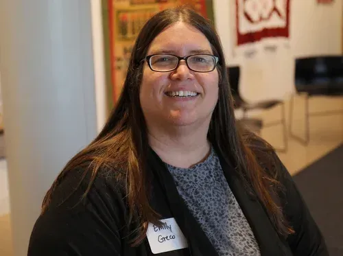 Smiling person with glasses and long brown hair in a black blazer, wearing a name badge, indoors.