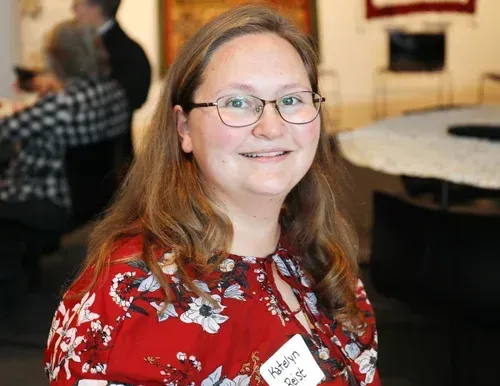Smiling person with glasses and long hair in a red floral shirt at a table in an indoor event space