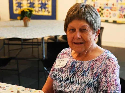 Smiling woman seated at a table in a decorated room, wearing a patterned top and name tag.