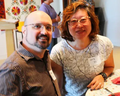 Two smiling people posing indoors at a table, one in glasses and a striped shirt, the other in a white patterned top.