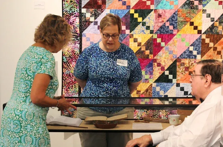 Three people discussing a colorful quilt display at a table.