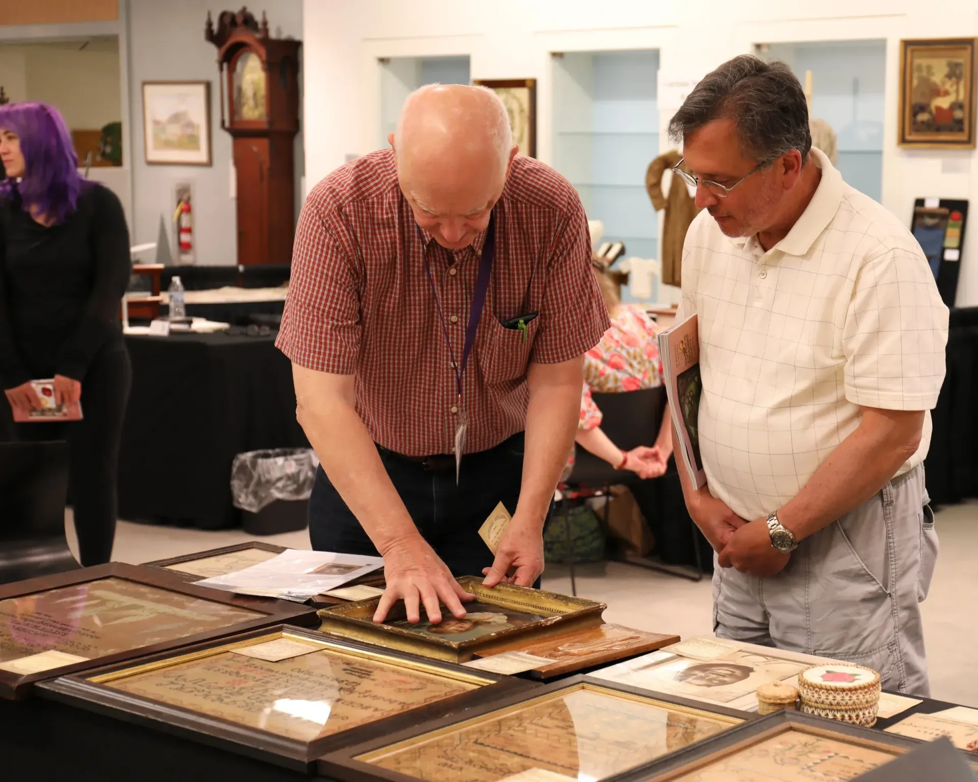 Two people examine framed artwork on a table in a gallery-like room.