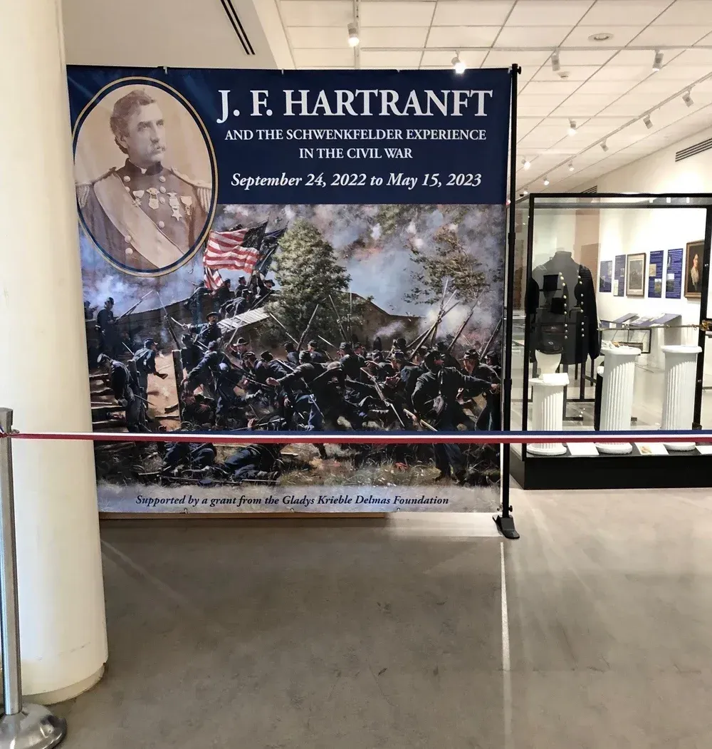 Museum exhibit banner for J.F. Hartman with historical portrait, dates, and crowd scene behind a barrier