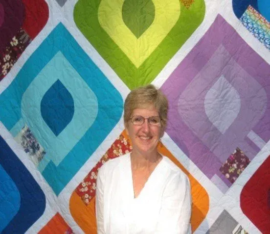 Smiling woman in white shirt standing before a colorful quilt with diamond patterns.