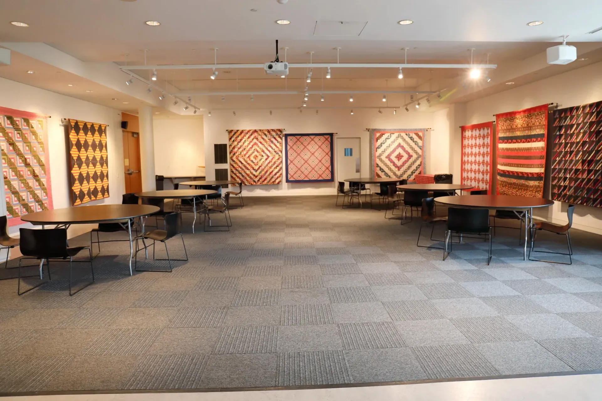 Wide gallery room with large colorful quilt displays on the walls and tables arranged across a carpeted floor