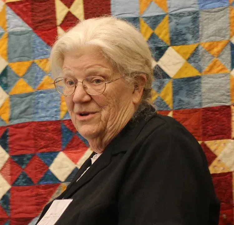 Elderly woman in glasses and black top standing before a colorful quilt backdrop