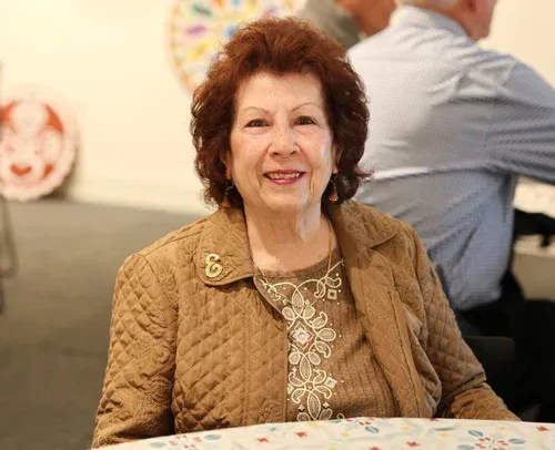 Smiling woman seated at a table in a warmly lit indoor gathering, wearing a tan jacket and lace top.