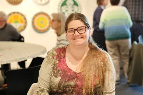 Smiling woman with glasses seated at a table in a bright indoor gathering space