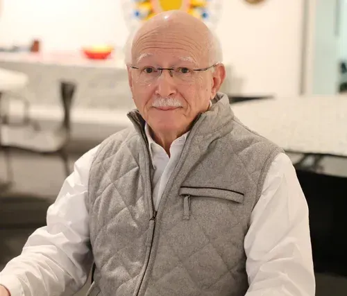 Man wearing glasses and a light gray quilted vest seated indoors against a blurred office background