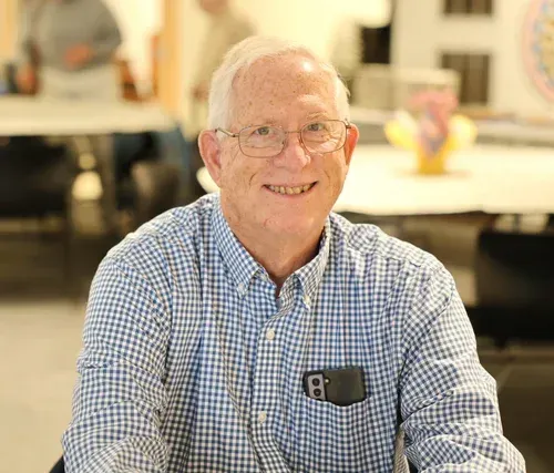 Smiling man in a blue checkered shirt seated indoors, with a blurred café-like background
