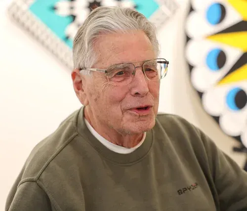 Older man in glasses and olive sweatshirt, seated indoors with abstract wall art behind him