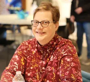 Smiling person in a red floral shirt and glasses seated indoors at a table