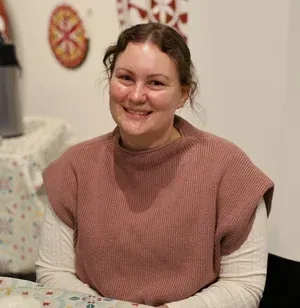 Smiling person in a pink sweater seated indoors against a light wall.