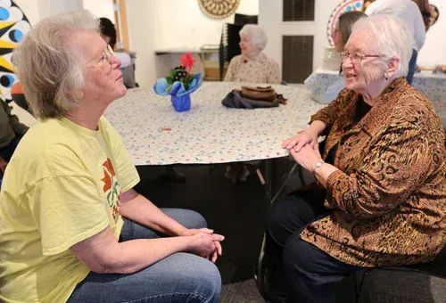 Two older adults chatting at a table in a bright dining room, with a blue vase of flowers between them.