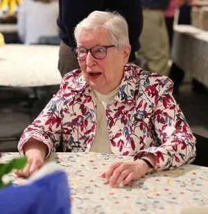 Elderly woman in glasses and floral jacket talking at a table in a social setting