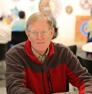 Person in a red and black jacket sitting indoors at a table, with blurred artwork in the background