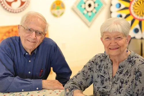 Two smiling people seated at a table with colorful wall art behind them.