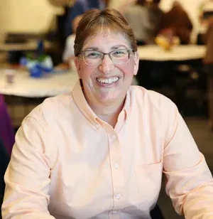 Smiling person in a light pink shirt seated at a table in a blurred indoor setting