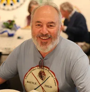 Smiling man in a gray T-shirt with a “Battle Field” graphic, seated indoors at a table.