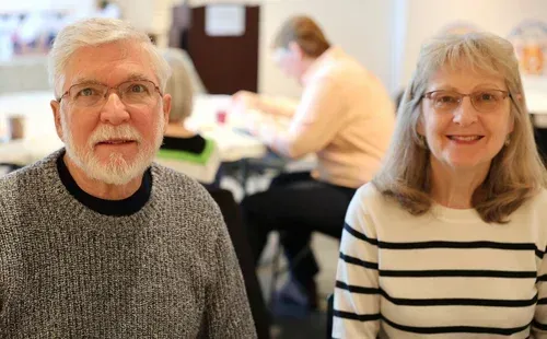 Two smiling people seated indoors, with a blurred person working in the background.