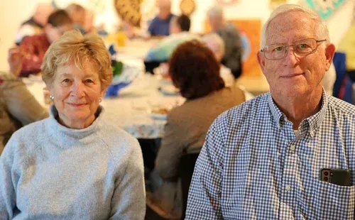 Two smiling people seated indoors at a social gathering, with blurred guests in the background