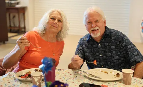 Two people smiling at a dining table with plates and cups, one holding a fork, in a bright room